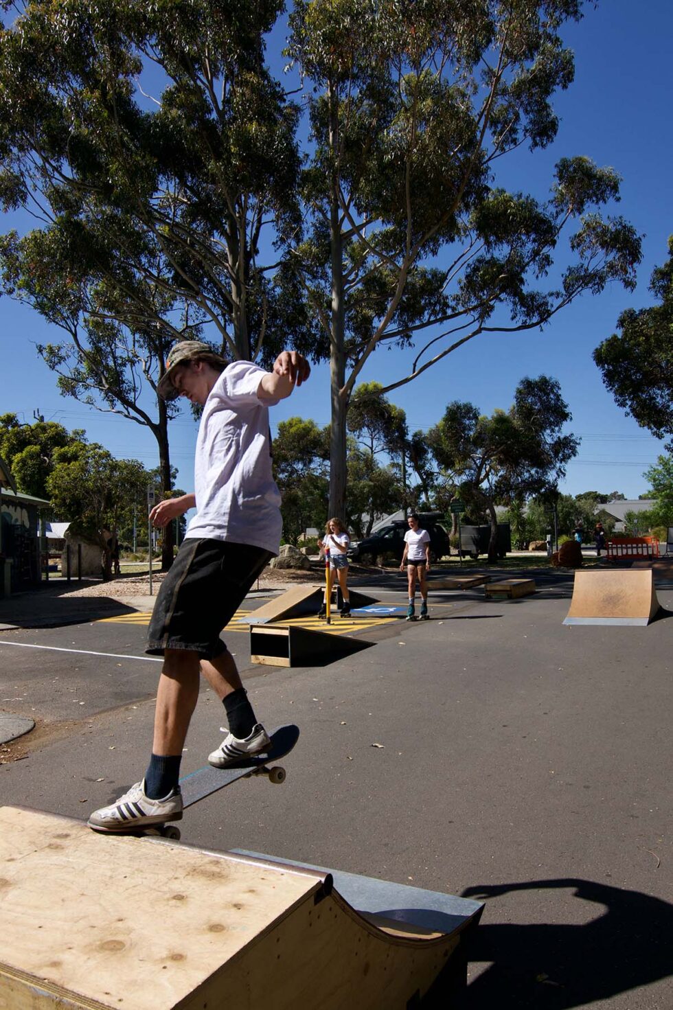 Concrete Club A skateboarder performs a trick on a ramp in an outdoor skate park surrounded by trees, under a clear sky. Other skateboarders are visible in the background.