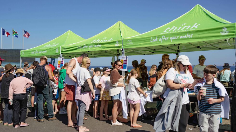 Concrete Club People stand in line under green marquees at an outdoor event on a sunny day. Some are holding food and drinks. Flags are visible in the background. The words Think, Look after your mates is written onthe marquees.