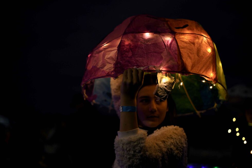 Concrete Club A person holds a colorful, illuminated umbrella shaped paper and cane hand-made prop, at night, casting a soft glow on their face.