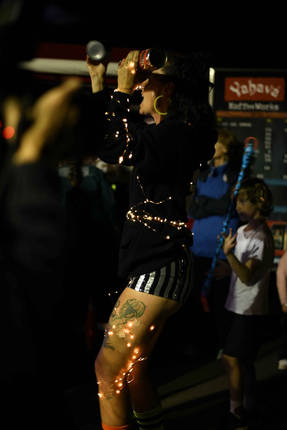Concrete Club Jacque dressed in dark clothing with fairy lights wrapped around their body holds two cans like musical shakers, while people play in the background.