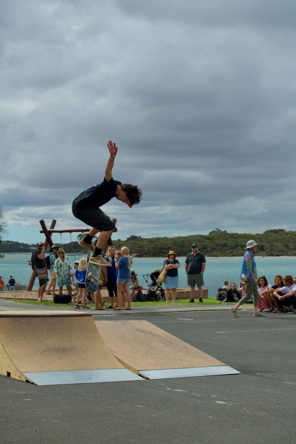 Concrete Club A skateboarder performs an airborne trick on a ramp near a body of water, with a crowd of spectators observing under a cloudy sky.