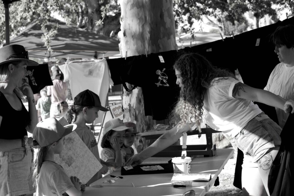 Concrete Club People gathered at an outdoor booth interact, with Kahleah and Elisha on the right handing items to children and adults who are observing or picking up posters. A row of shirts is displayed in the background.