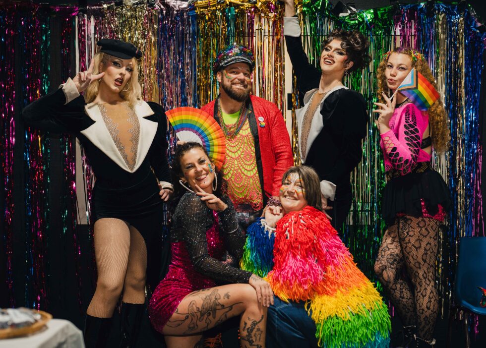 Concrete Club A group of six people dressed in colorful, flamboyant outfits pose in front of a shiny, tinsel backdrop, celebrating with rainbow-themed accessories. Concrete Club Pride.