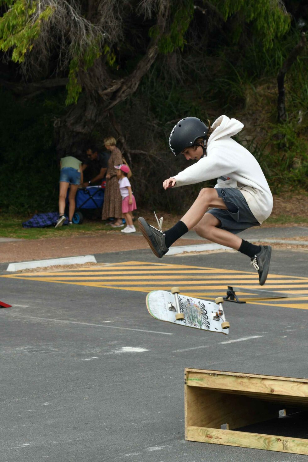 Concrete Club A person in a white hoodie and helmet is performing a skateboard trick in the air while wearing knee socks and shorts. A wooden ramp and skateboard are visible below. People are in the background.